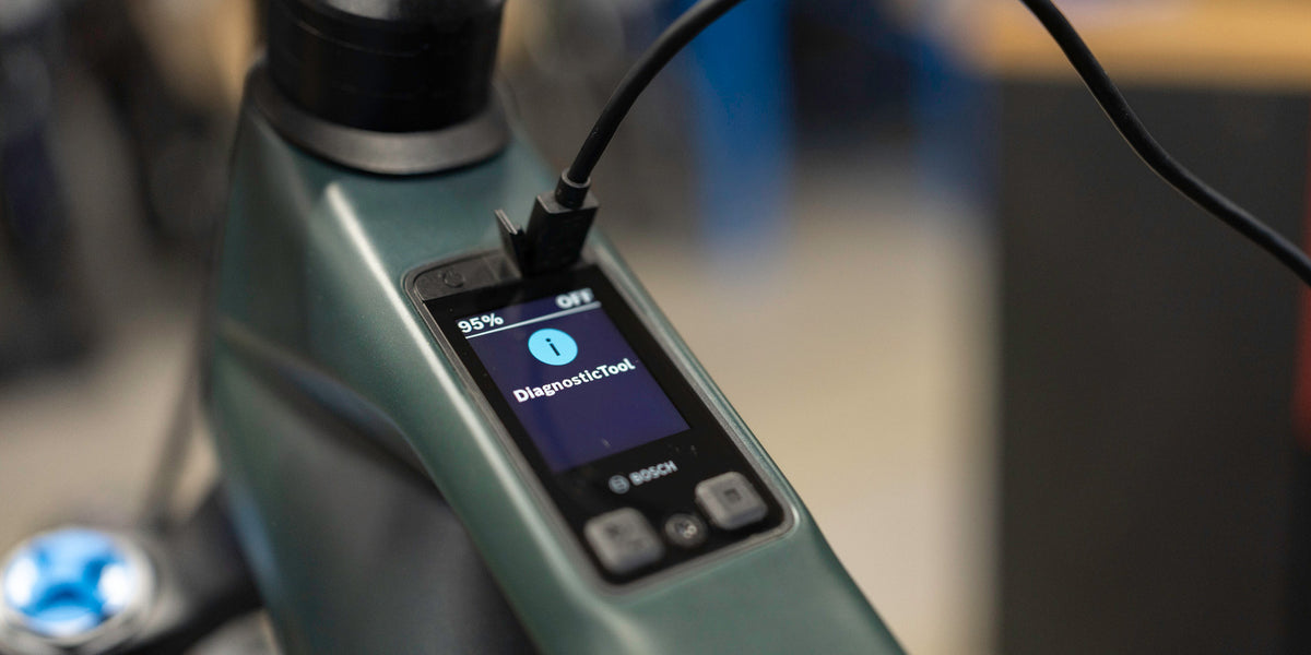 A close-up of a Bosch handheld diagnostic tool plugged into a green and black electric bike, showing its screen on.